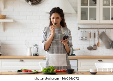 Smiling Young Woman In Apron Stand In Modern Kitchen Preparing Salad Use Smartphone Read Recipe, Happy Millennial Female Cooking Making Dinner Or Lunch Chatting Browsing On Modern Cellphone