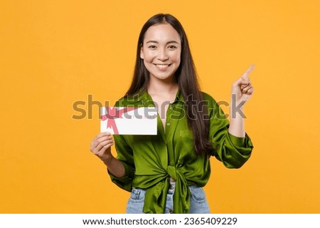 Smiling young woman of Asian ethnicity wear green shirt hold in hand gift certificate pointing indicate on workspace area copy space mock up isolated on bright yellow color background studio portrait