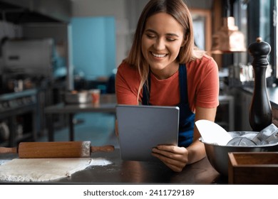 Smiling young woman baker using digital tablet at work in the pastry kitchen. Beautiful happy female baker looking at fintech device while working in the bakery. - Powered by Shutterstock