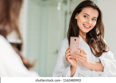 smiling young woman in bathrobe taking selfie with smartphone at mirror in bathroom  - Powered by Shutterstock