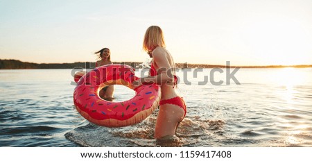 Smiling young woman in a bikini holding a swim ring and splashing her friend with water while enjoying an afternoon at a lake