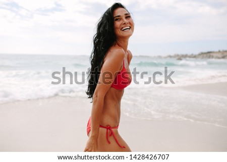Smiling young woman in bikini walking on the sea shore. Beautiful female enjoying summer vacation on the beach.