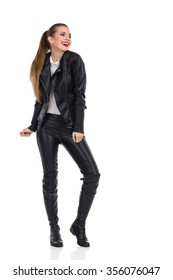 Smiling Young Woman In Black Leather Trousers, Jacket And Boots Posing And Looking Away. Full Length Studio Shot Isolated On White.