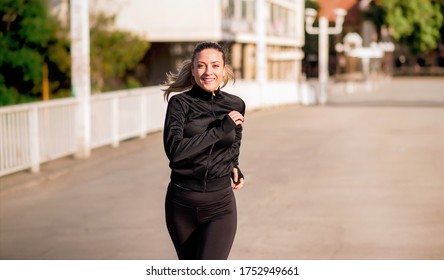 Smiling Young Woman In Black Sports Gear Running Across A Walking Bridge And Looking Towards The Camera. Healthy Morning Run. 