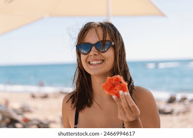 A smiling young woman in a black swimsuit stands under an umbrella on a sandy beach, holding a juicy slice of watermelon with the ocean and blue sky behind her. - Powered by Shutterstock