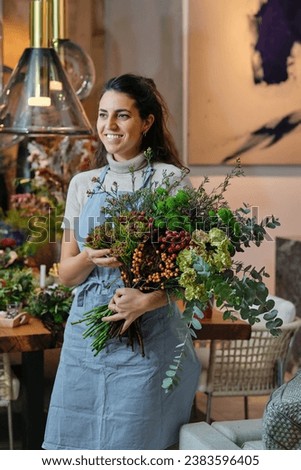 Smiling young woman in blue apron touching arranged flower bouquet and looking away while working in floral shop on blurred background