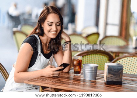 Smiling young woman with blue eyes sitting on urban cafe using smart phone. Girl with brown wavy hairstyle wearing white denim dress. Lifestyle concept.