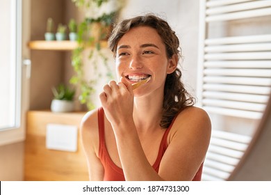 Joven sonriente cepillando dientes en el baño. Chica feliz mirando en espejo mientras usa cepillo de dientes ecológico con pasta dentífrica blanquecina. Chica de belleza en el baño limpiando dientes en la mañana.