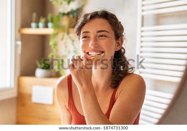 Smiling young woman brushing teeth in bathroom.\
Happy girl looking in mirror while using ecological toothbrush with\
whitening toothpaste. Beauty girl in bathroom cleaning teeth in the\
morning time.