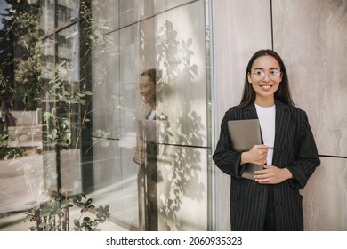 Smiling Young Woman In Casual Clothes Standing In Office Not In Full Growth. Businesswoman In Glasses With Laptop In Hand, Looking At Camera.