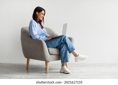 Smiling young woman in casual wear working online, sitting in armchair and using laptop against white studio wall, copy space. Cheerful Caucasian lady surfing internet on portable pc - Shutterstock ID 2021617133