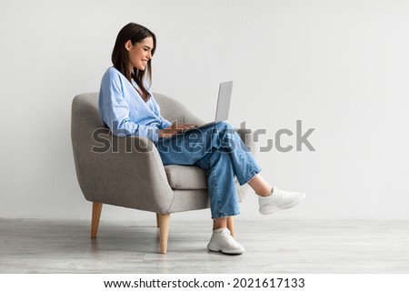 Smiling young woman in casual wear working online, sitting in armchair and using laptop against white studio wall, copy space. Cheerful Caucasian lady surfing internet on portable pc