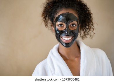 Smiling Young Woman With Charcoal Face Mud Looking At Camera Isolated On Beige Background With Copy Space. Portrait Of African Woman Wearing Bath Robe Getting Black Purifying Peeling Mask At Spa.