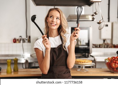 Smiling young woman chef cook in apron standing at the kitchen, holding ladle and whisk - Powered by Shutterstock