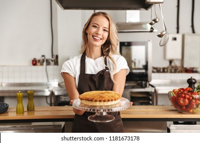 Smiling Young Woman Chef Cook In Apron Standing At The Kitchen, Showing Tasty Pie