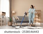 Smiling young woman cleaning rug with cordless vacuum cleaner in living room