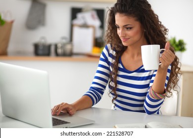 Smiling Young Woman With Coffee Cup And Laptop In The Kitchen At Home