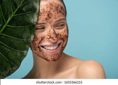 smiling young woman with coffee scrub on face, isolated on blue with leaf