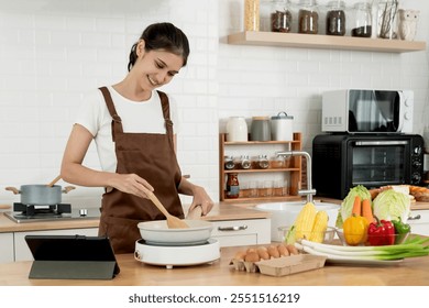 Smiling Young Woman Cooking in Modern Kitchen, Wearing Apron and Using Tablet for Recipe, Surrounded by Fresh Vegetables and Ingredients, Healthy Lifestyle and Home Cooking Concept - Powered by Shutterstock