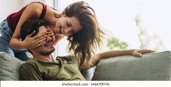 Smiling young woman covering her partner's eyes sitting on sofa. Young woman surprising man in the living room at home. - Powered by Shutterstock