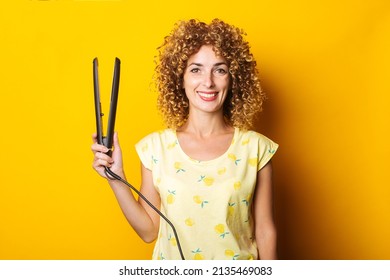 Smiling Young Woman With Curly Hair Holding Hair Straightener On Yellow Background.