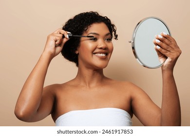 A smiling young woman with curly hair is applying mascara to her eyelashes as she looks into a handheld mirror. She is wrapped in a white towel or cloth - Powered by Shutterstock