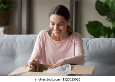 Smiling Young Woman Customer Open Parcel Cardboard Box Sit On Sofa At Home, Happy Girl Consumer Unpacking Carton Package Looking Inside Receive Good Purchase Post Mail Shipping Delivery Concept