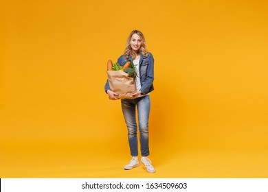 Smiling Young Woman In Denim Clothes Isolated On Yellow Orange Wall Background. Delivery Service From Shop Or Restaurant Concept. Hold Brown Craft Paper Bag For Takeaway Mock Up With Food Products