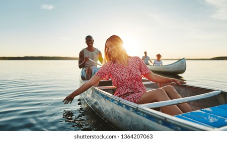 Smiling Young Woman Dipping Her Hand In Lake Water While Paddling A Canoe With Her Boyfriend And Friends On A Late Summer Afternoon