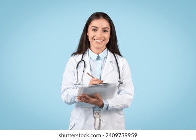 Smiling young woman doctor in white lab coat writing medical notes using digital tablet, representing modern healthcare, blue background - Powered by Shutterstock