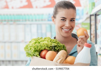 Smiling young woman doing grocery shopping at the supermarket, she is holding a bag with fresh vegetables, bread and milk