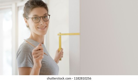 Smiling young woman doing a home makeover, she is measuring a wall using a folding ruler, DIY and house renovation concept