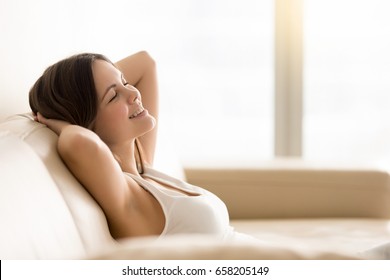 Smiling young woman dreaming about summer leisure while sitting with closed eyes and hands behind head on sofa at home. Happy lady relaxing in hotel room during vacation. Pleasant memories concept - Shutterstock ID 658205149
