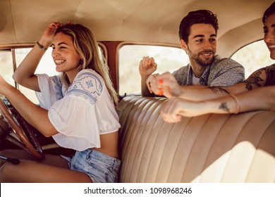 Smiling Young Woman Driving Car With Her Friends Sitting In The Backseat. Friends Going On A Road Trip In Old Car.