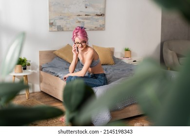 Smiling Young Woman With Dyed Hair Sitting On Bed While Talking On Mobile Phone