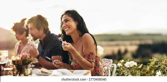 Smiling young woman eating food and talking with friends during a dinner party outdoors. Woman enjoying having food with friends at a party.