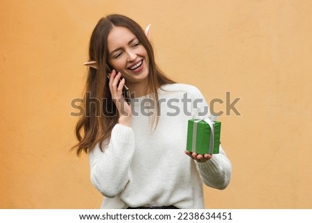 Smiling young woman with elf ears talking on mobile phone and holding green gift box in hand, against yellow background 