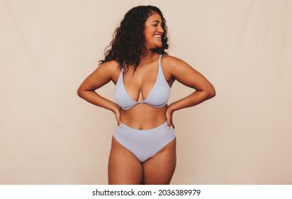 Smiling young woman embracing her natural body and curves in a studio. Happy young woman wearing underwear and feeling positive about her body. Young woman standing alone against a studio background.