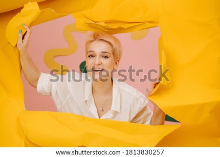 Smiling young woman emerging through a hole in a big sheet of cramped yellow paper, tearing it up. She has short dyed blond hair and stars eye makeup.
