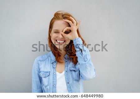 Smiling young woman encircling her eye with her fingers as she grins happily at the camera over a grey background