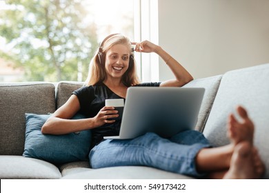 Smiling young woman enjoying a cup of coffee while relaxing with her laptop in living room. Female sitting on sofa with headphones watching movie on laptop.
