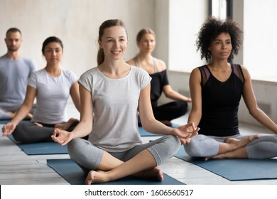 Smiling young woman enjoying practicing yoga at group lesson, sitting in Easy Seat pose on mat, diverse people doing Sukhasana exercise, stress relief, working out in modern yoga center club