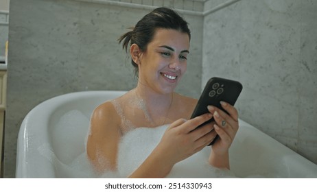 A smiling young woman enjoys a bubble bath while using her smartphone in a modern tiled bathroom. - Powered by Shutterstock