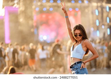 Smiling young woman enjoys summer music festival on beach, dancing among crowd. Hand up with peace sign, trendy sunglasses and casual festival wear. Sunset lights add warm, vibrant feel to scene. - Powered by Shutterstock