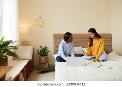 Smiling young woman explaining difficult topic to friend when they are sitting on bed in dormitory