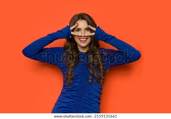Smiling young woman in fitted\
cornflower blue dress with long sleeves looks through her fingers.\
Three quarter length studio shot against an orange\
background.