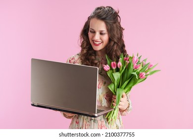 smiling young woman in floral dress with tulips bouquet using laptop isolated on pink.
