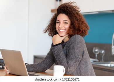A smiling young woman with frizzy hair working from home on laptop in kitchen.