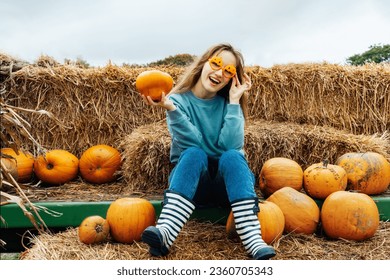 Smiling young woman in fun glasses sitting on straw bales and holding pumpkin. Selecting best one for Thanksgiving and Halloween holidays decoration on agriculture farm. Autumn fall festive mood.