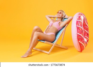 Smiling young woman girl in pink striped swimsuit sit on deck chair isolated on yellow background studio. People summer vacation rest lifestyle concept. Mock up copy space. Holding hands behind head